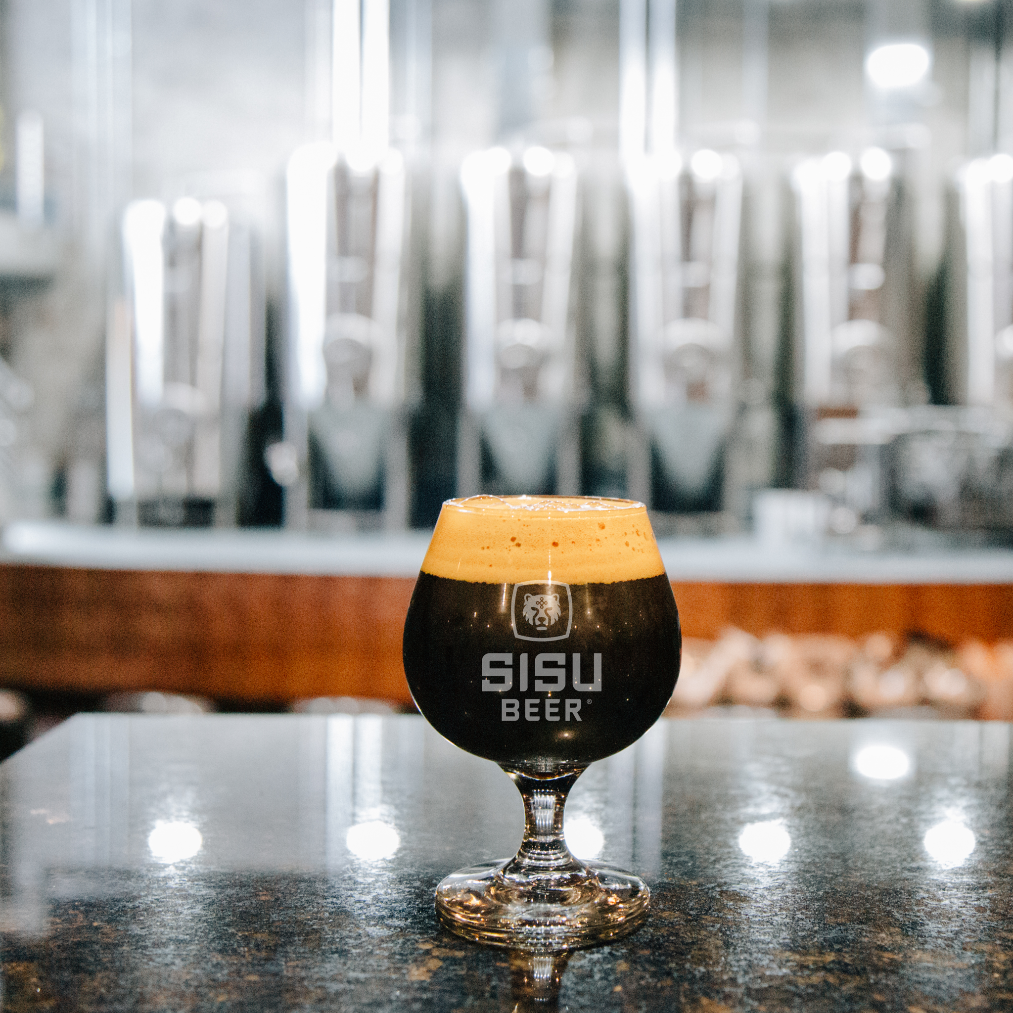 Snifter of dark Sisu stout on the bar with brewery tanks behind